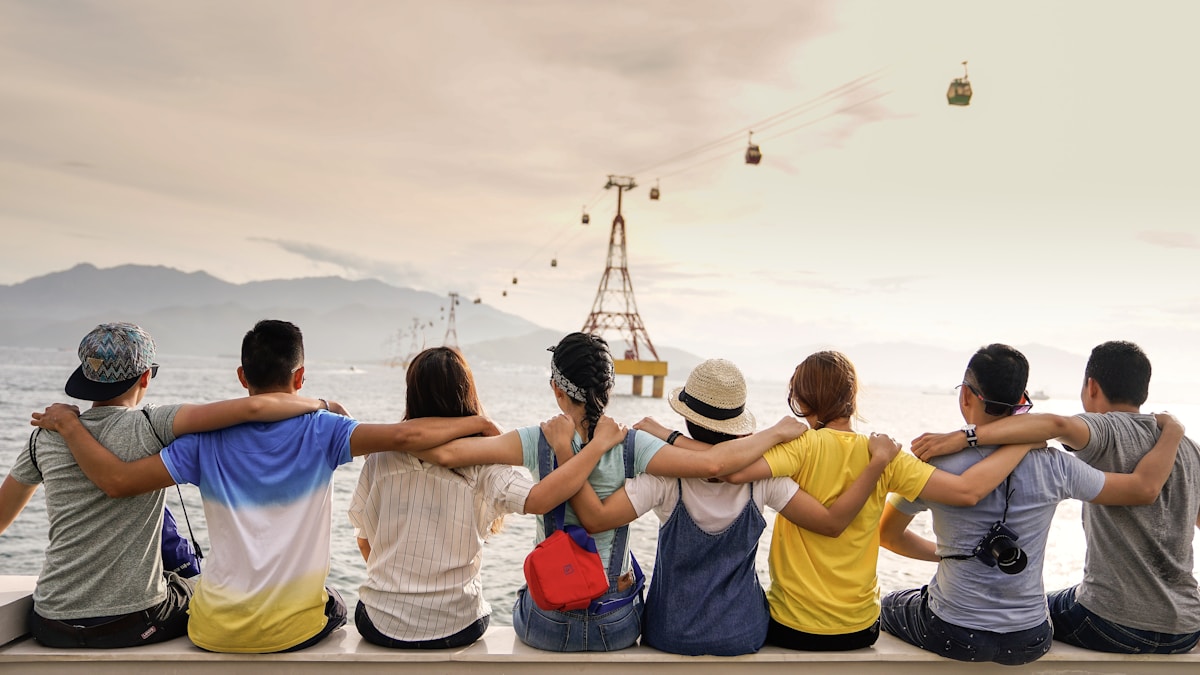 Eight friends sitting side by side with arms around each other, facing cable cars over water and distant mountains